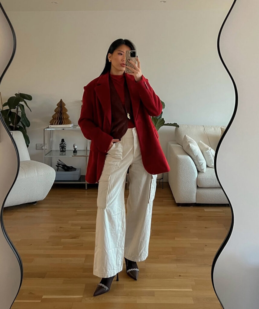 Mirror selfie of woman wearing a red coat over a burgundy top with cream cargo trousers and brown pointed heels in a minimalist living room.