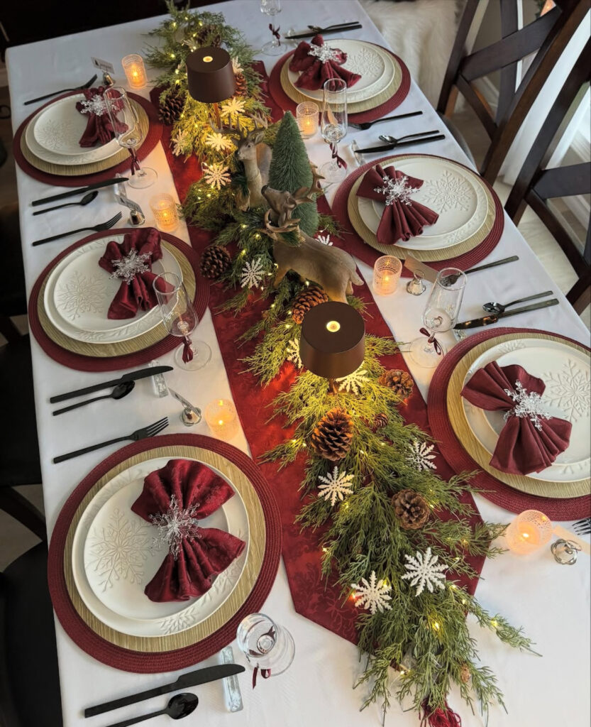 Long table with red runner, lit evergreen garland, deer figurines, pinecones, and burgundy bow napkins.
