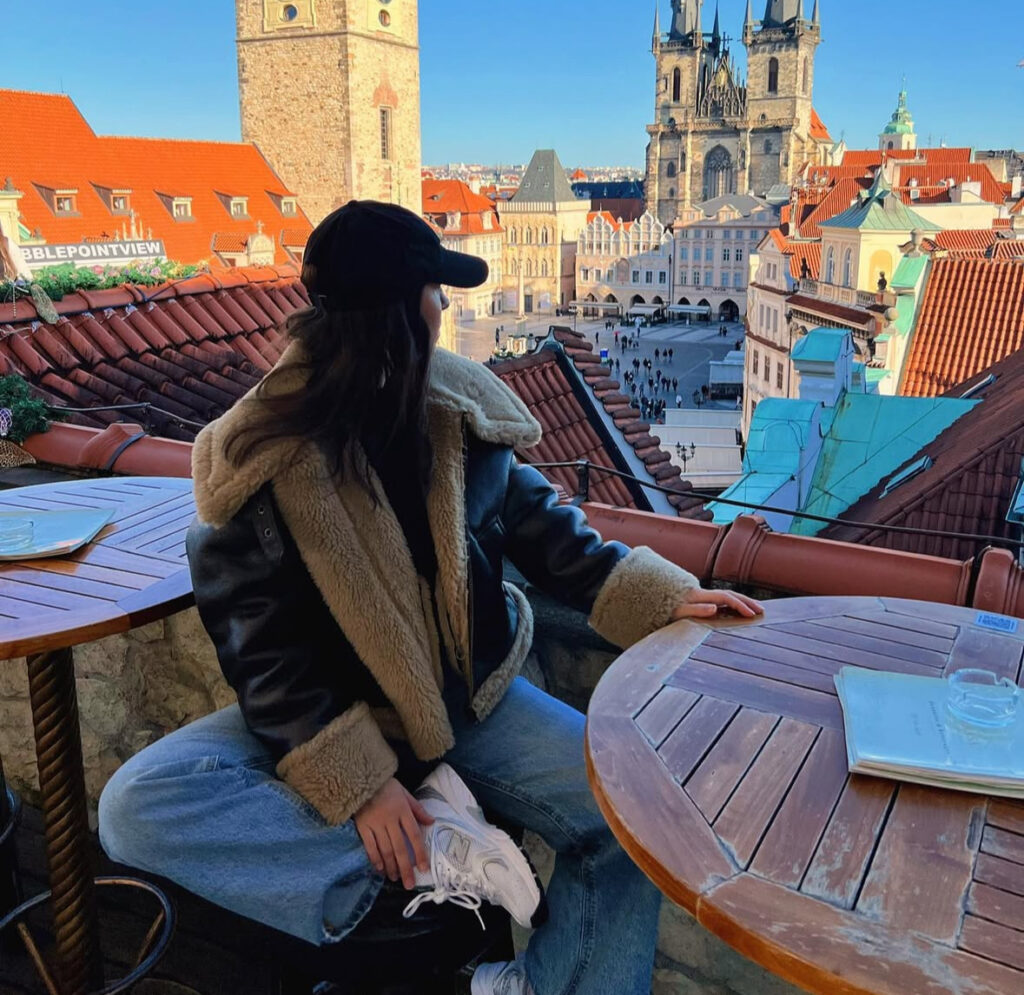 Shearling-trim jacket with straight-leg jeans, clean white sneakers, and a cap on a rooftop with skyline.