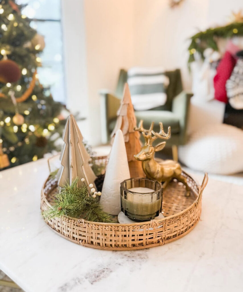 Round woven tray on marble table with ceramic and wood trees, gold deer figurine, greenery, and a small candle.