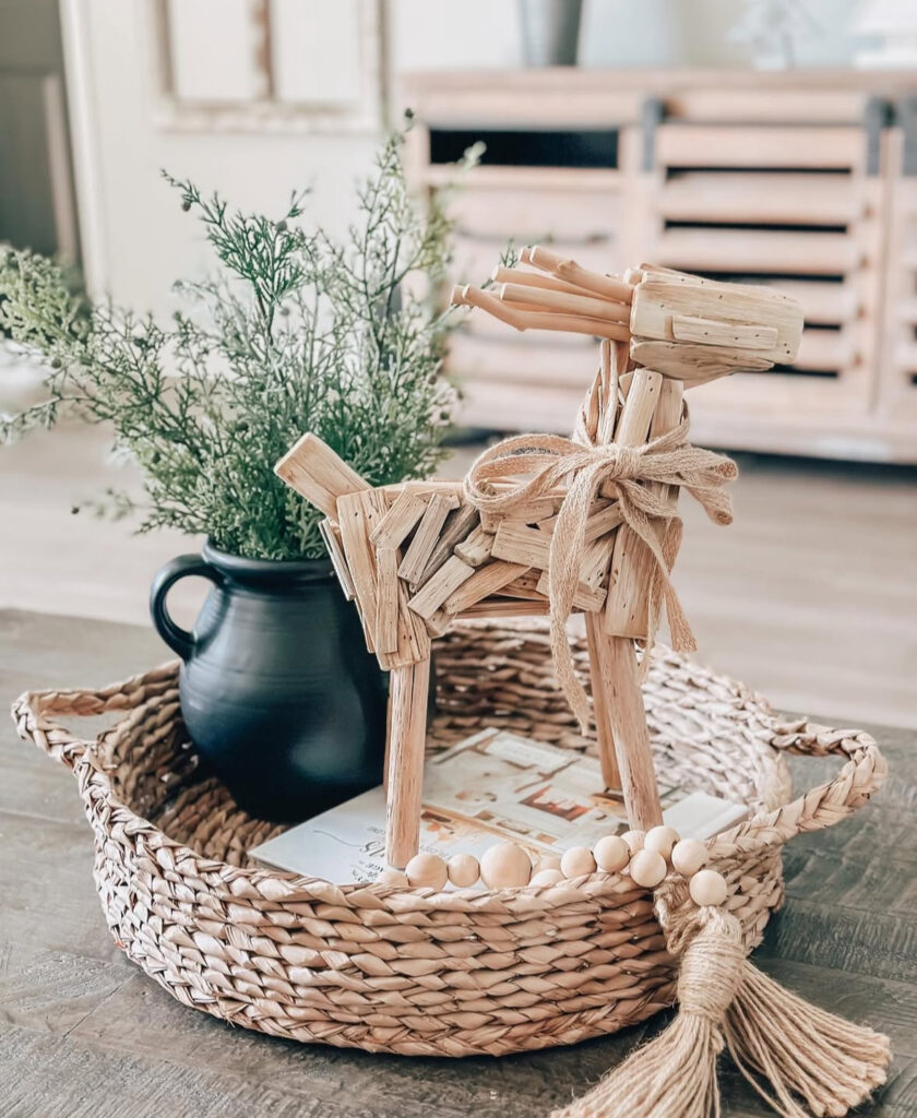 Woven basket tray with a black jug of greenery, wood-bead garland, and a straw reindeer figurine on a rustic coffee table.