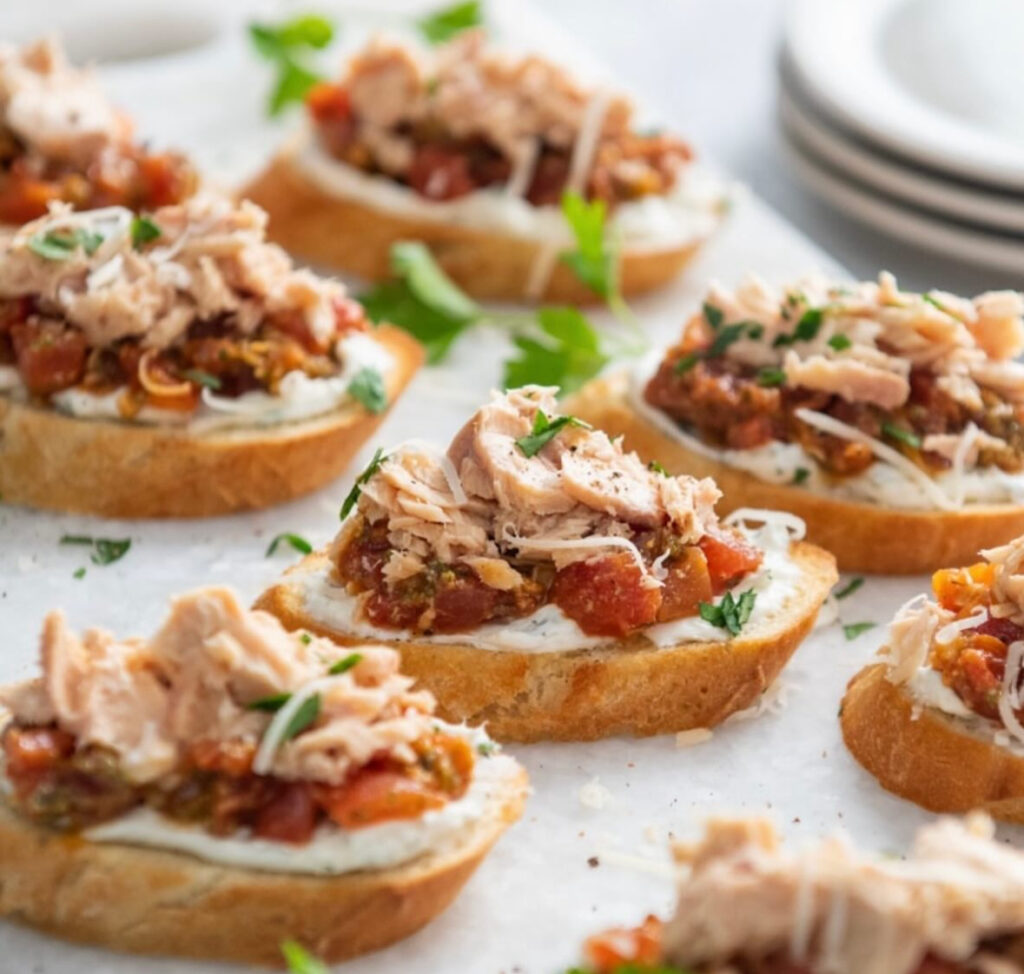 Baguette slices layered with herbed spread, tomato relish, and flaky pink salmon with parsley.
