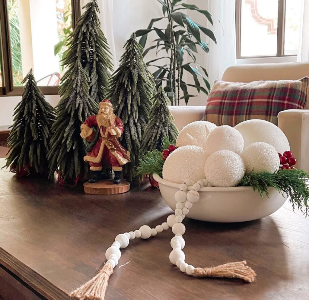 Cluster of faux pine trees with vintage Santa figurine beside a bowl of white foam “snowballs,” greenery, and bead garland on a wood table.