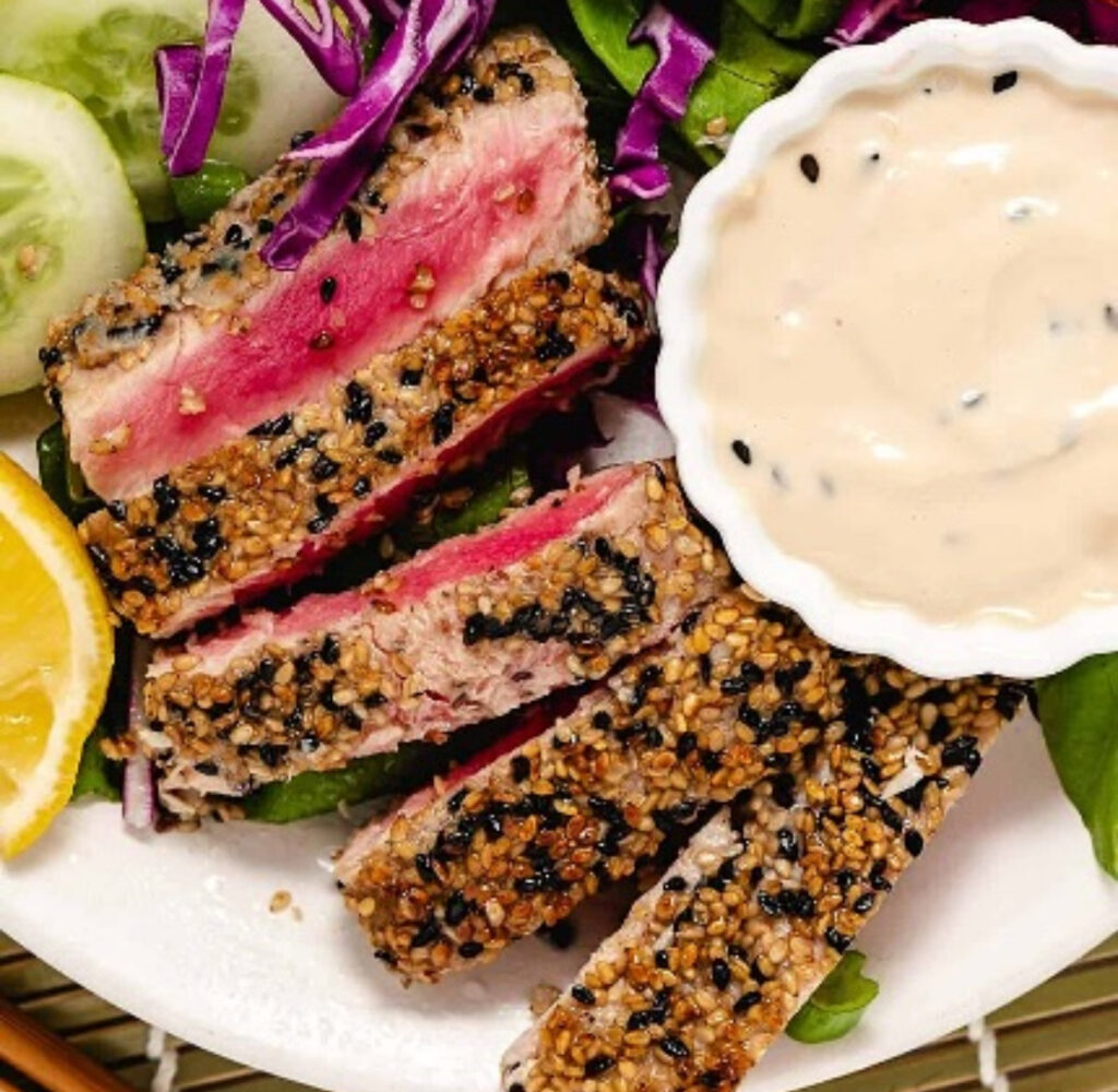 Sliced ahi tuna with black and white sesame crust, pink center, lemon wedge, and creamy dipping sauce.