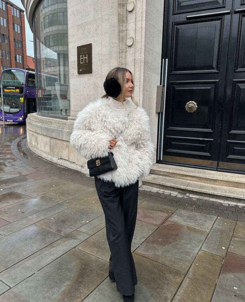 Shaggy ivory jacket with tailored black trousers and a small shoulder bag near city buildings.
