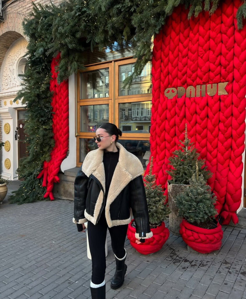 Woman in oversized black shearling aviator jacket, black outfit and chunky boots walking past festive red knit decor and small trees.