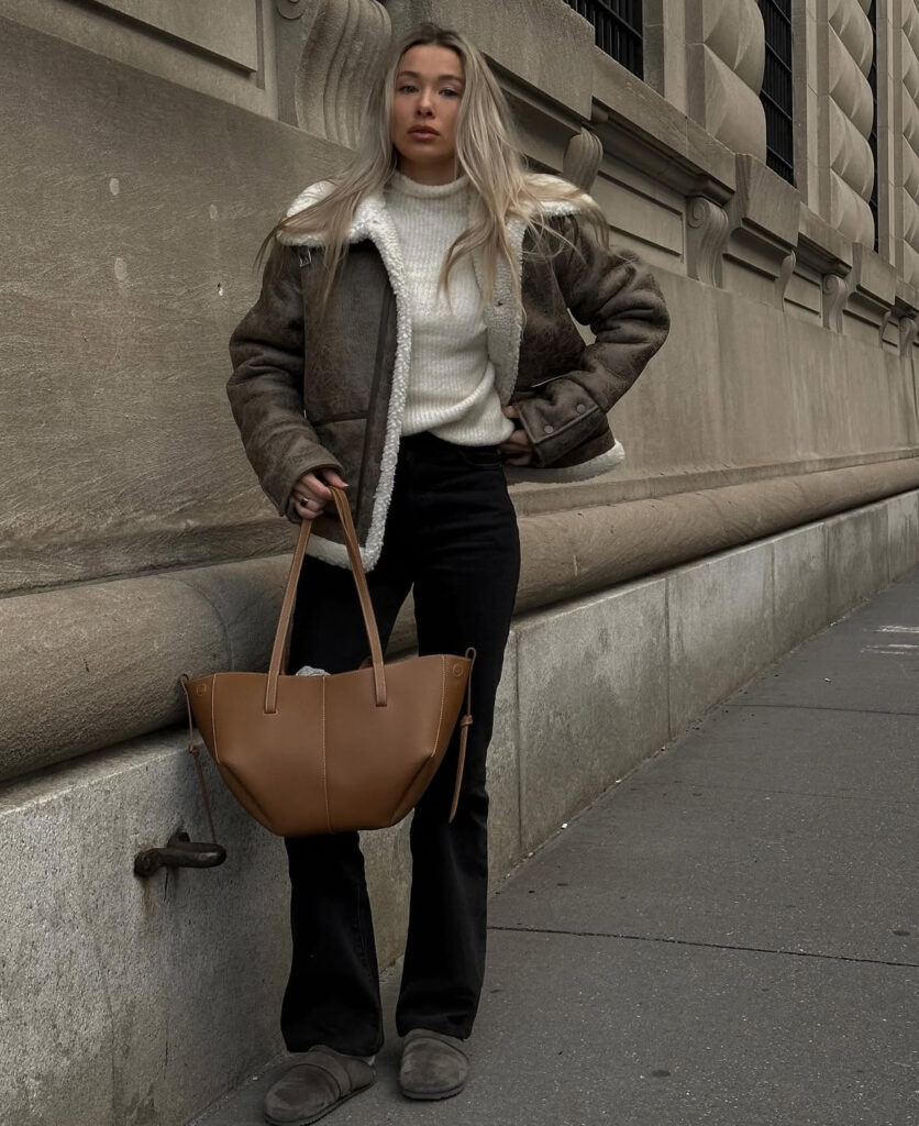 Aviator shearling over chunky knit with black flares, clogs, and a brown tote, walking in the city.
