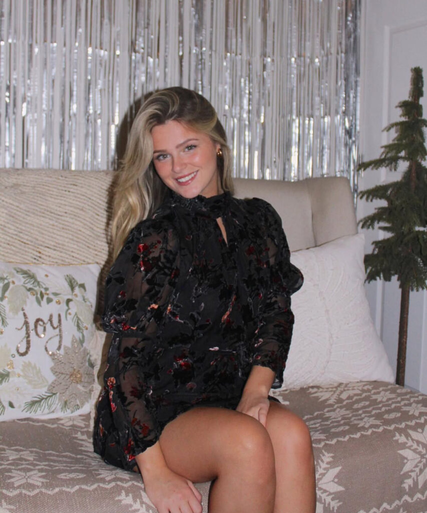 Woman sitting and smiling in a black floral sheer long-sleeve mini dress in a festive room with silver fringe backdrop.