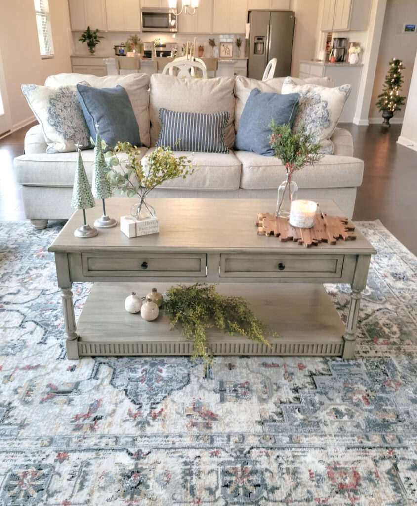 Neutral sofa with blue pillows; rectangular table styled with ceramic trees on stands, a small vase of greens, and a glowing candle on a wood trivet.