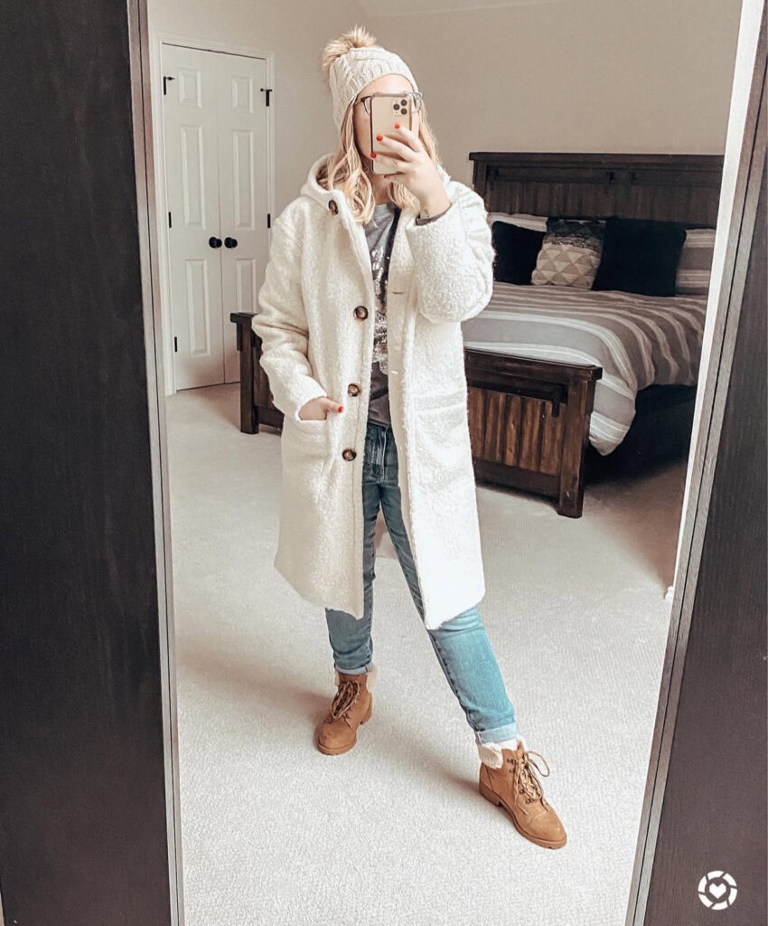 Mirror selfie of woman in a white sherpa coat with hood, knit beanie, cuffed jeans and tan winter boots in a bedroom.