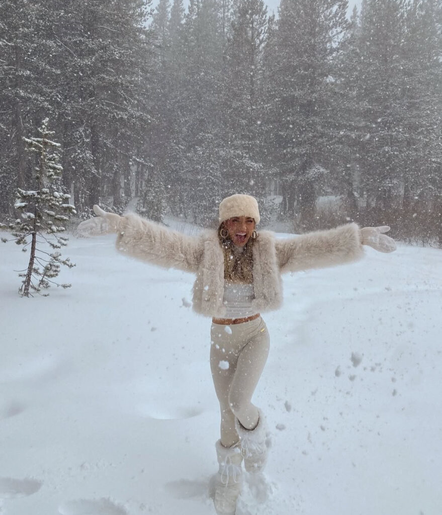 Fuzzy white jacket, matching hat, and moon boots; sparkly details in a snowy street scene.