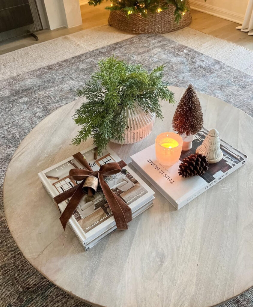 Round light wood table with stacked books tied in velvet, ribbed vase of greenery, pinecone, mini trees, and a small candle.