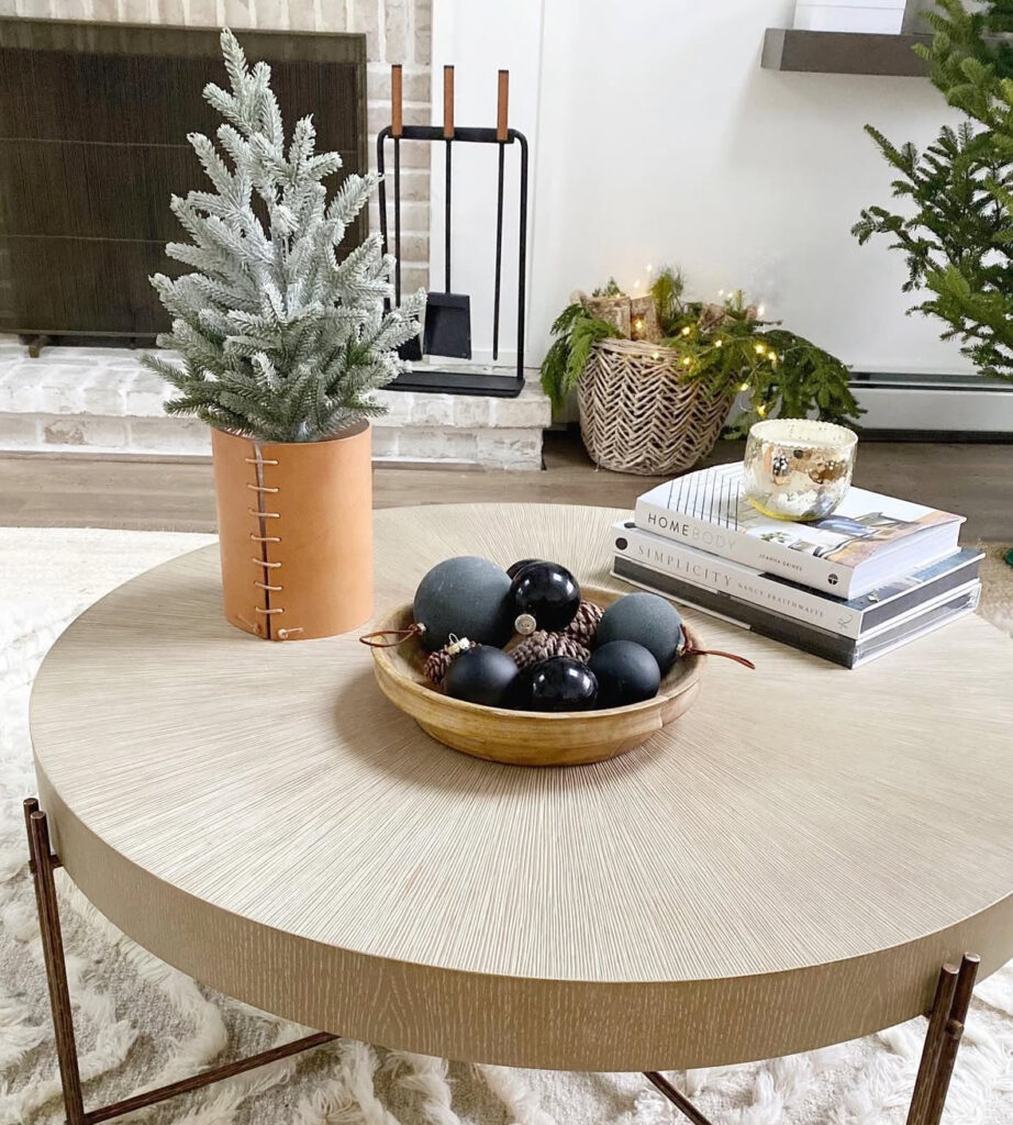 Light round table with leather-wrapped vase holding a mini fir, wood bowl of black ornaments, stacked books, and a mercury jar candle.