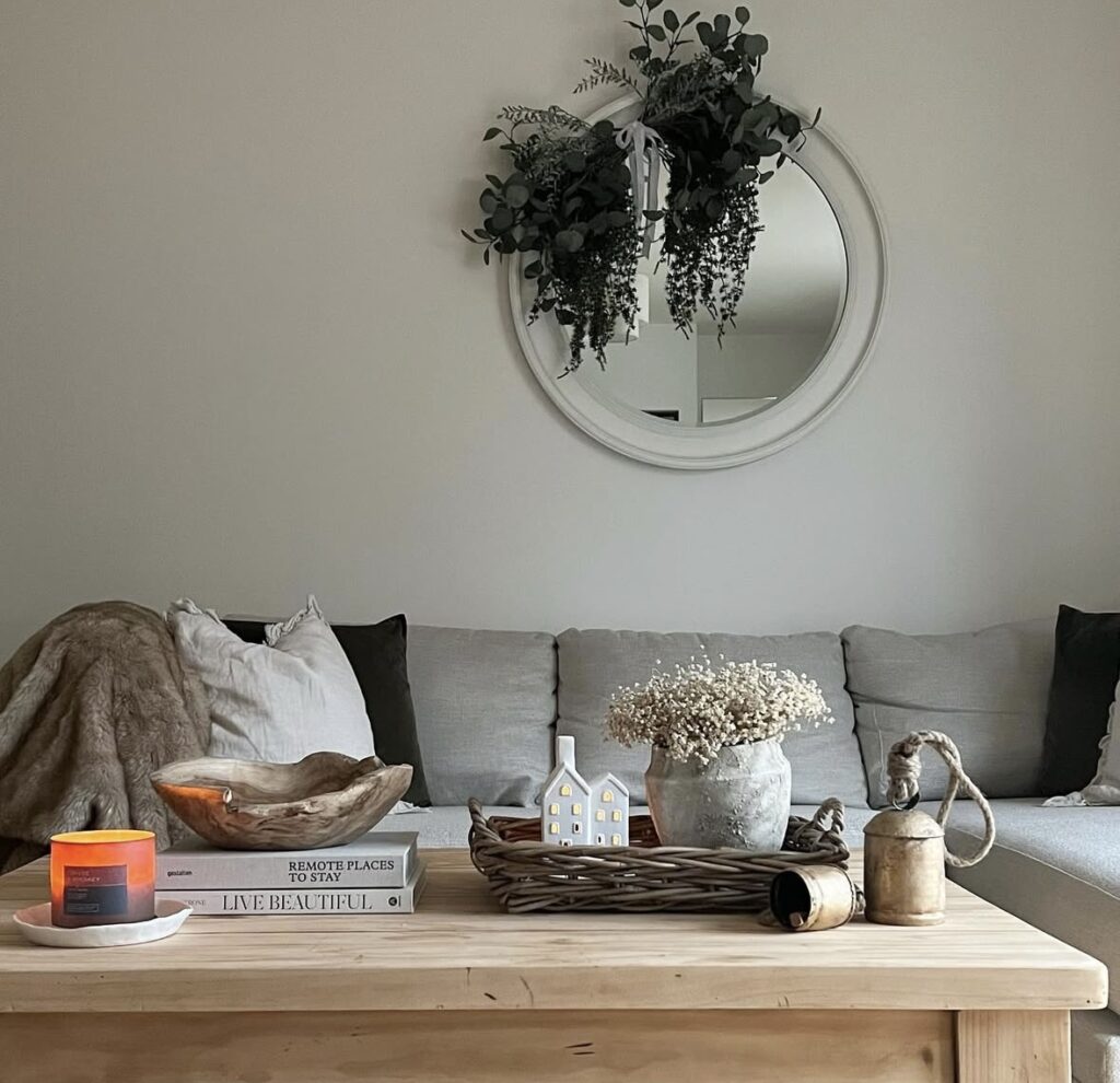 Neutral living room with a round mirror and hanging greens; wood coffee table holds a woven tray, dried blooms, and a tiny lit ceramic house.