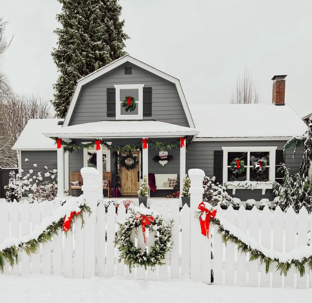 Snowy gray cottage with garland, red bows, and wreaths on a white picket fence.