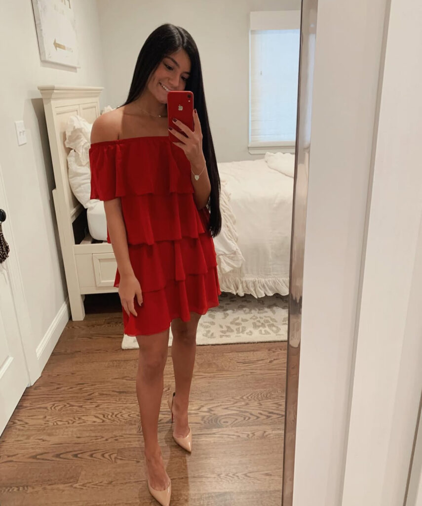 Mirror selfie of woman in tiered red off-shoulder mini dress with nude pointed pumps in a bright bedroom.