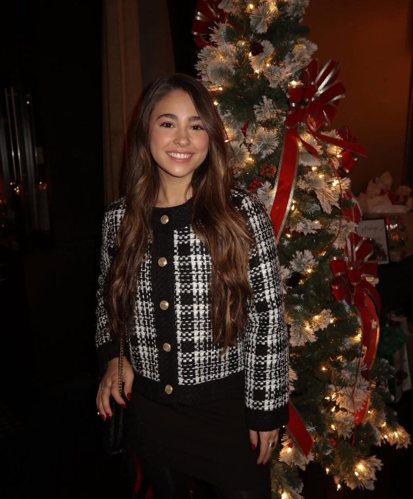 Woman in black-and-white tweed jacket with gold buttons over a black dress, smiling next to a lit Christmas tree.