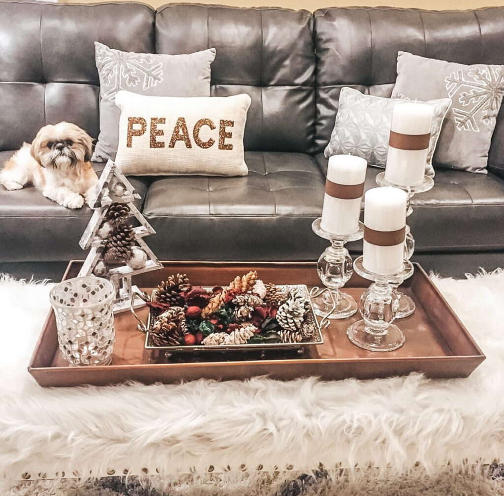 Copper tray with glass pillar candle holders, metallic pinecones, and a small acrylic tree on a faux-fur covered bench; dog on leather sofa behind.