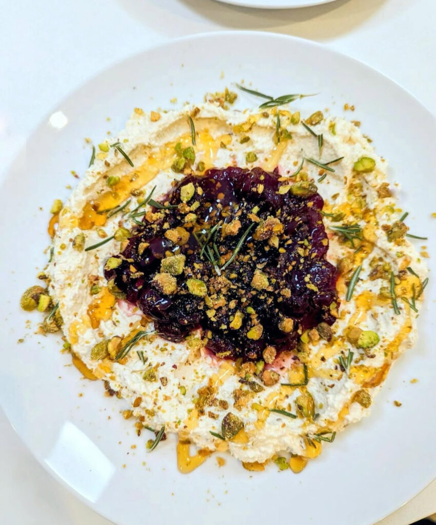 Wide white plate of whipped feta topped with cranberry compote, crushed pistachios, rosemary, and honey drizzle.