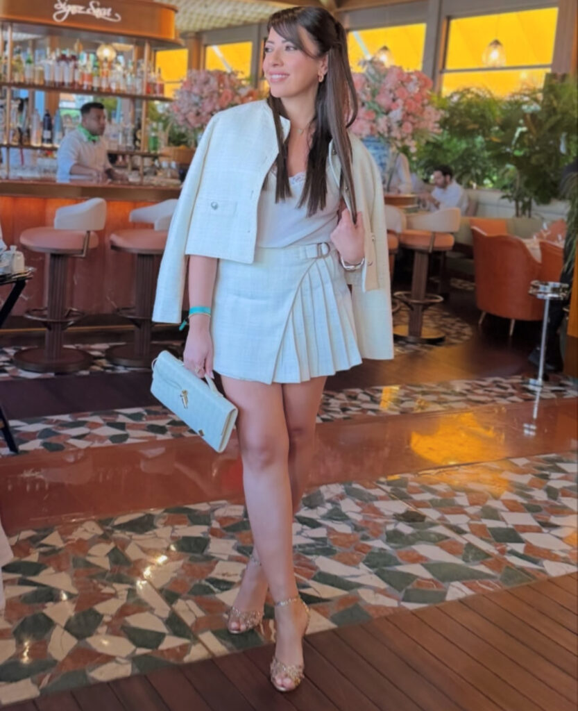 Woman in winter-white tweed skirt set with jacket over shoulders, sparkly heels and clutch inside a chic restaurant.