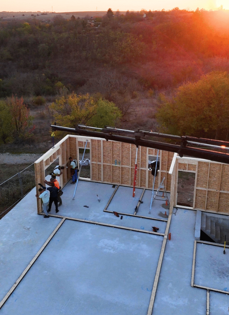 Crew lifting a wall panel by crane at sunset during first floor framing on a concrete slab.