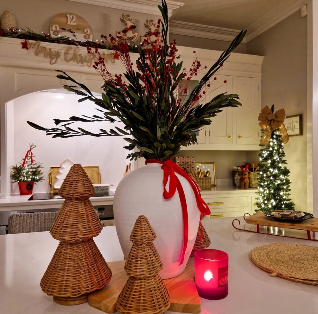 Evening island with large white vase and red ribbon, woven cone trees, and red candle; lit mini tree behind.