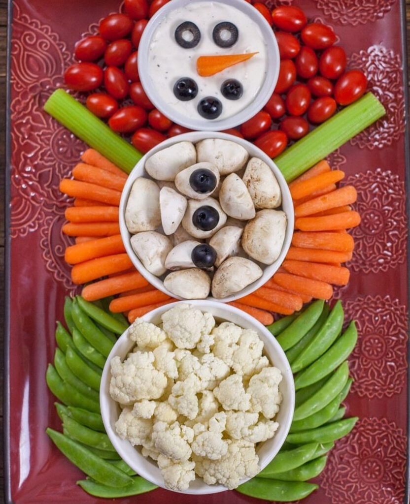 Snowman veggie tray made from three bowls of dip stacked vertically with peppercorn buttons, carrot nose, and veggies arranged around.