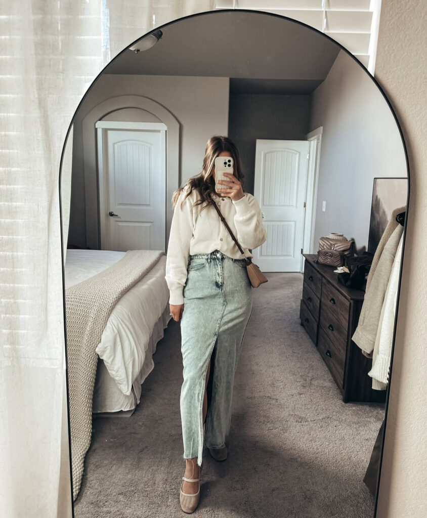 Mirror selfie of an ivory cardigan tucked into a light denim maxi skirt with a front slit, beige Mary-Jane flats, and a neutral crossbody.