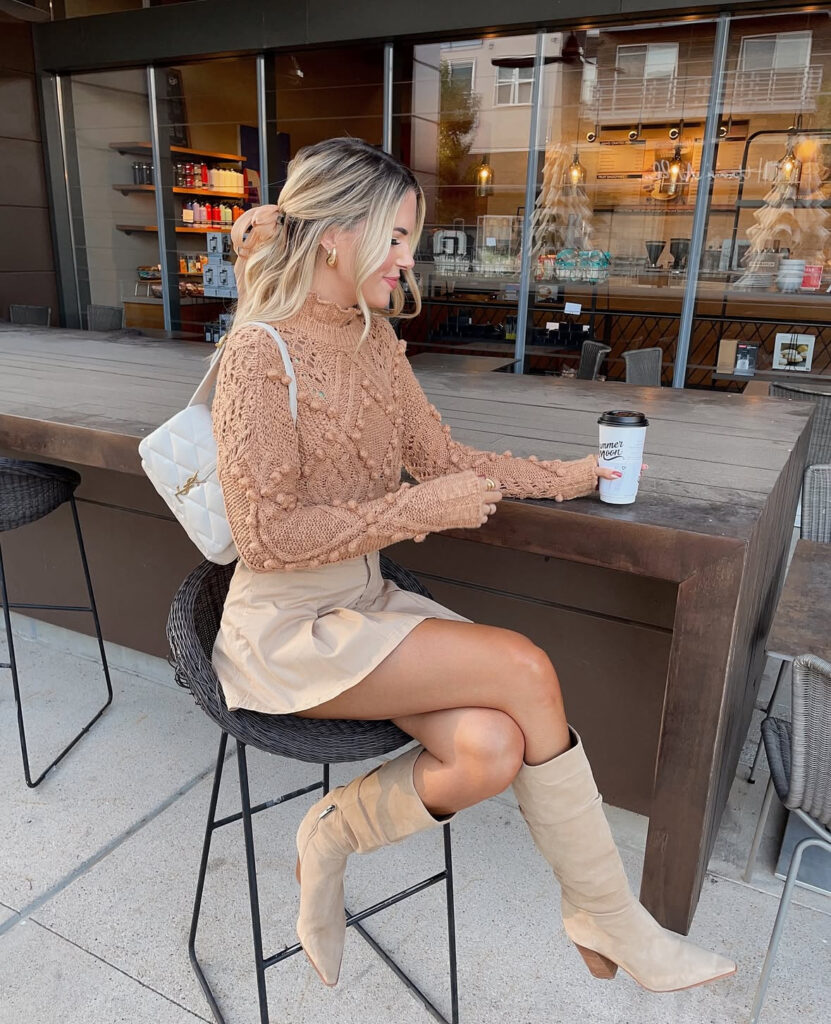 Woman at a café wearing a cinnamon textured sweater, beige mini skirt, and tan slouchy knee boots with a white quilted backpack, holding coffee.