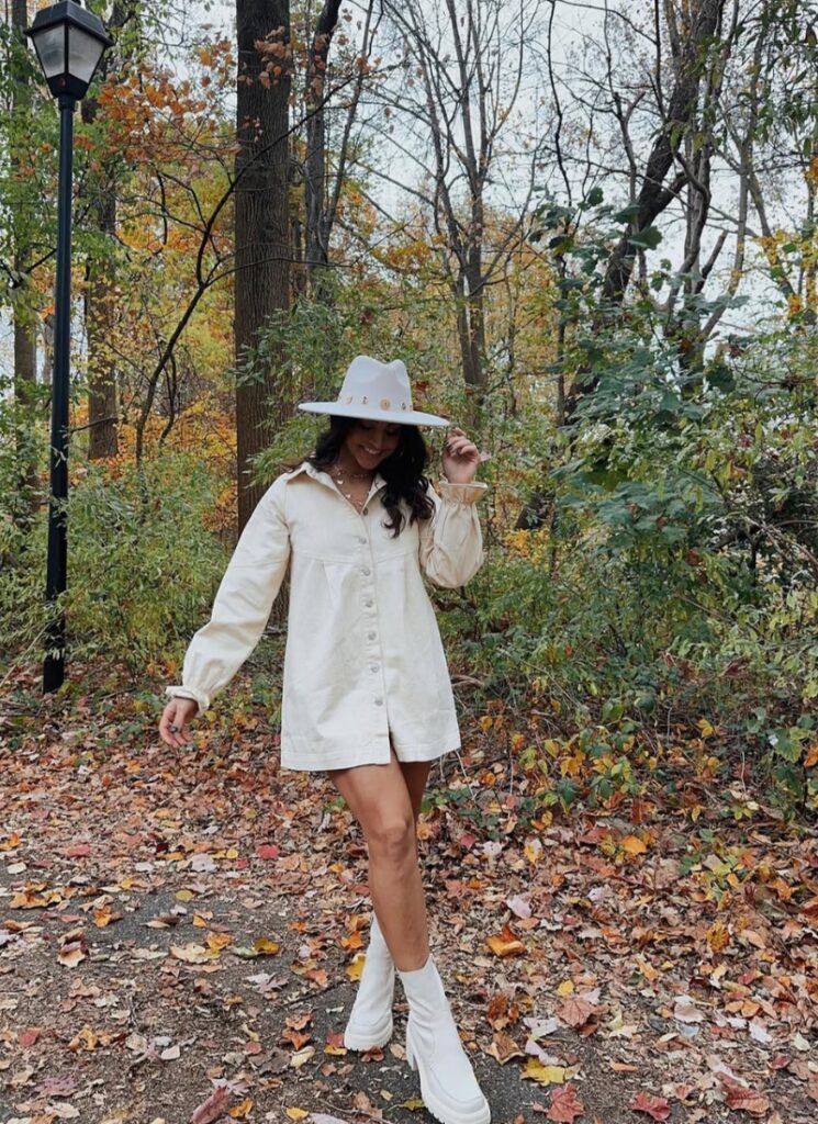 Woman walking through fall leaves in a cream buttoned shirt dress, white chunky boots, and a wide-brim hat.