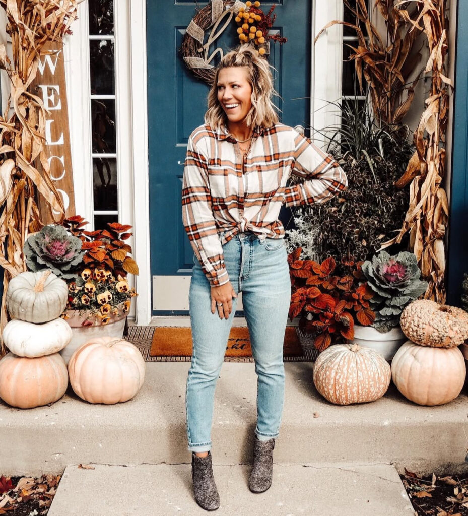 Woman on a pumpkin-filled porch wearing a cinnamon-cream plaid shirt knotted at the waist, light-wash jeans, and gray ankle boots.