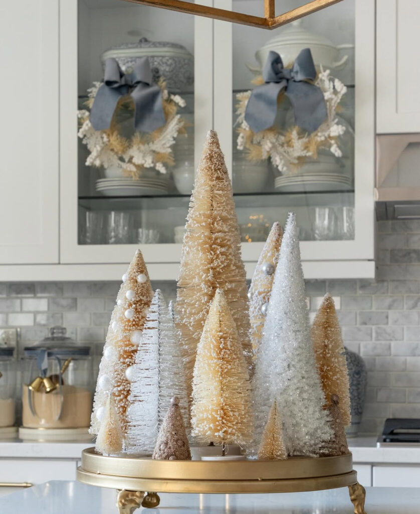 Gold-footed tray filled with champagne and white bottle-brush trees; glass cabinets with wreaths behind.
