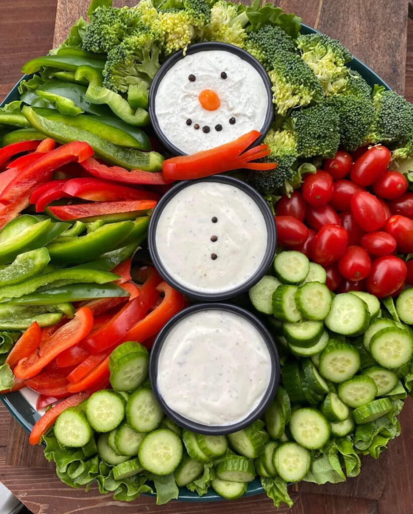 Cauliflower snowman on a clear plate with black olive eyes and mouth, long carrot nose, cucumber borders, and red pepper scarf.