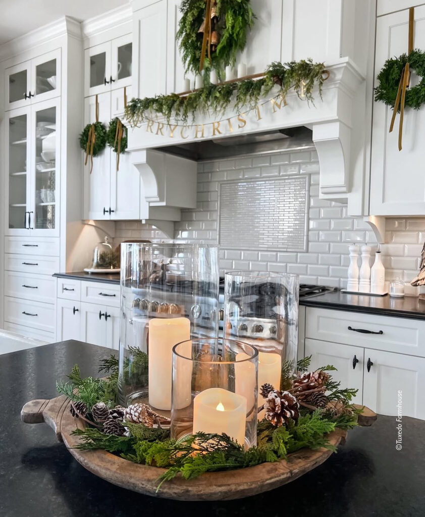 White kitchen with dough bowl centerpiece of glass hurricanes, candles, and greenery on the island.