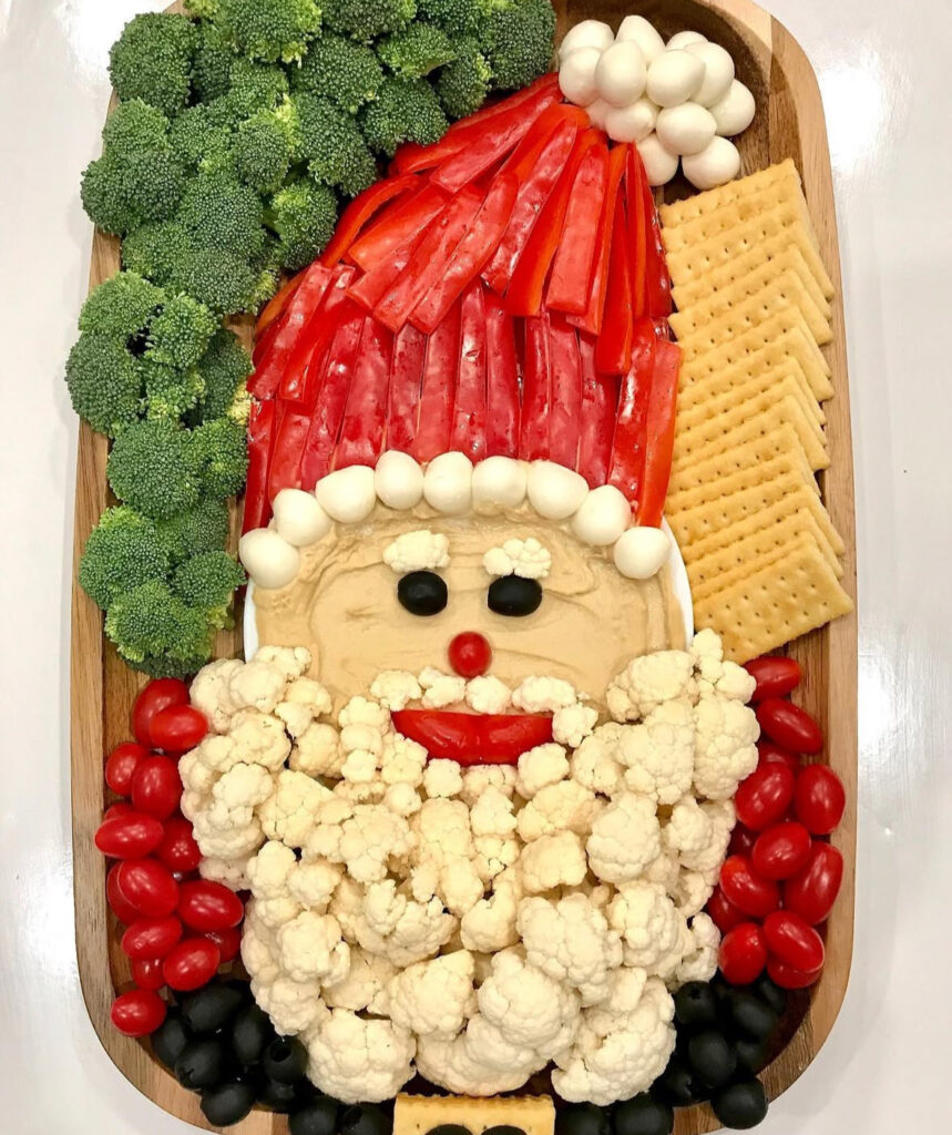 Hummus Santa platter with a red pepper hat, mozzarella pom-pom, cauliflower beard, crackers, olives, tomatoes, and broccoli.