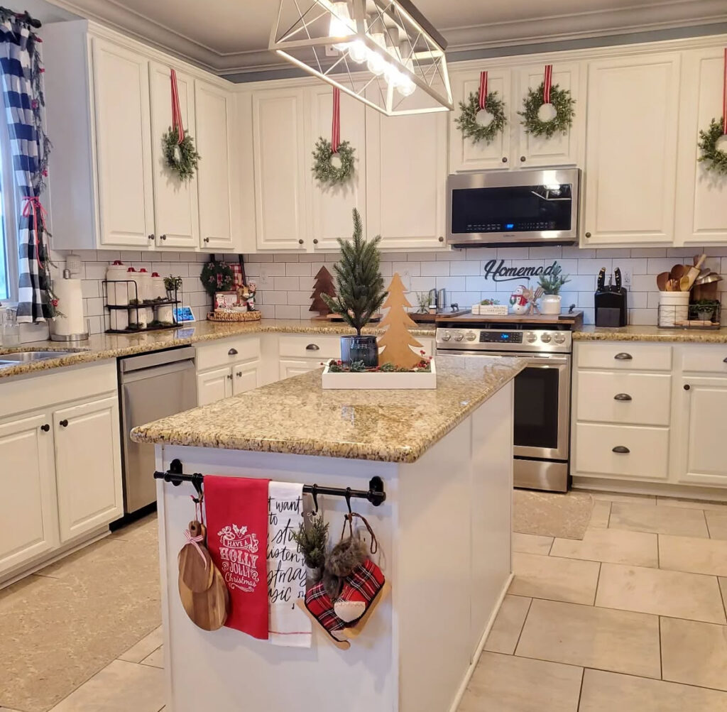 Neutral island with tray holding mini tree and wooden tree cutouts; cabinets hung with red-ribbon wreaths.