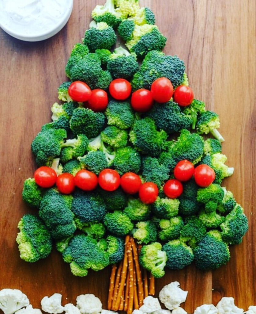 Minimalist broccoli tree on a wooden board with two rows of cherry tomatoes as garland and a pretzel-stick trunk, ranch dip at the corner.