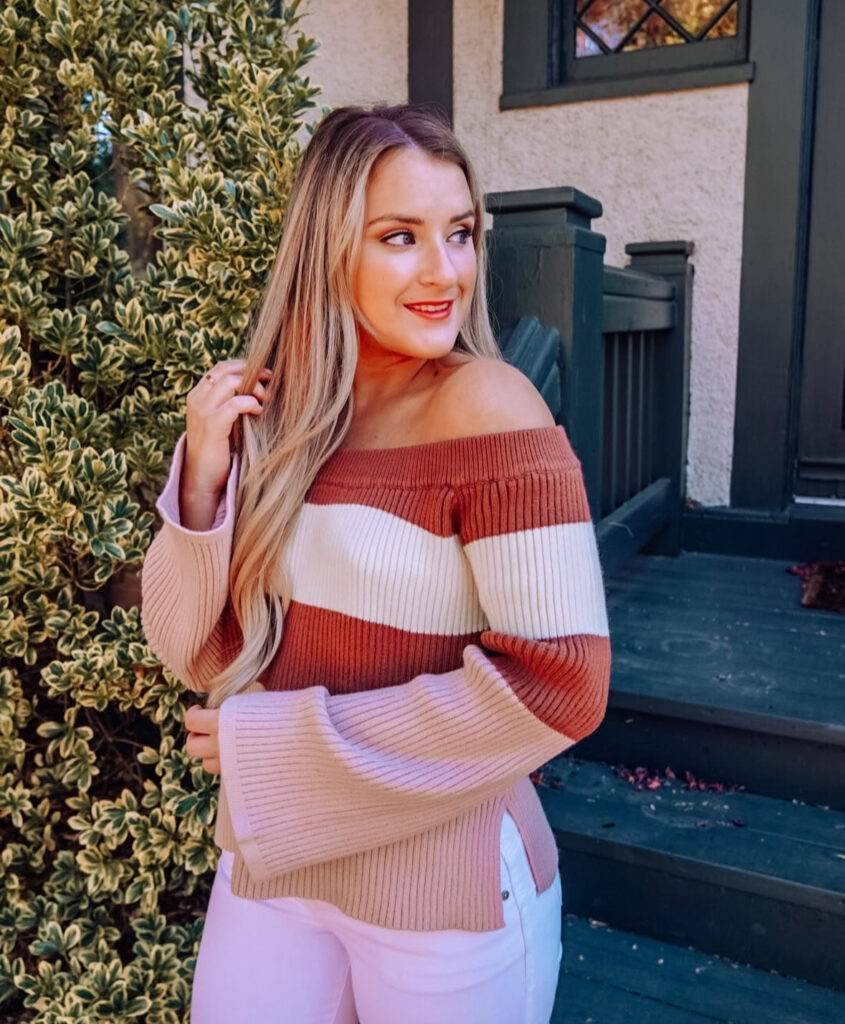 Close-up of an off-the-shoulder ribbed sweater in rust, cream, and blush stripes paired with white jeans.