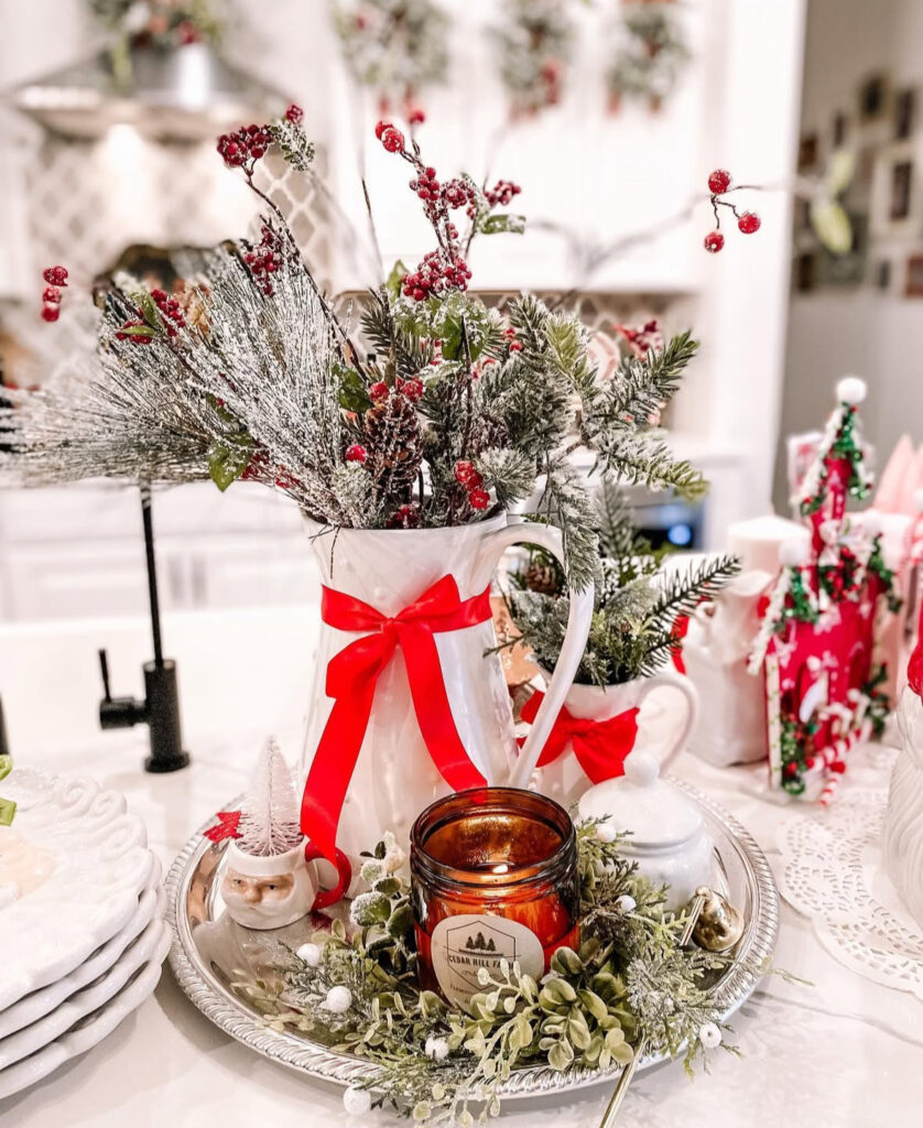 Silver tray with white pitcher of frosted greens tied with red ribbon and amber candle on island.
