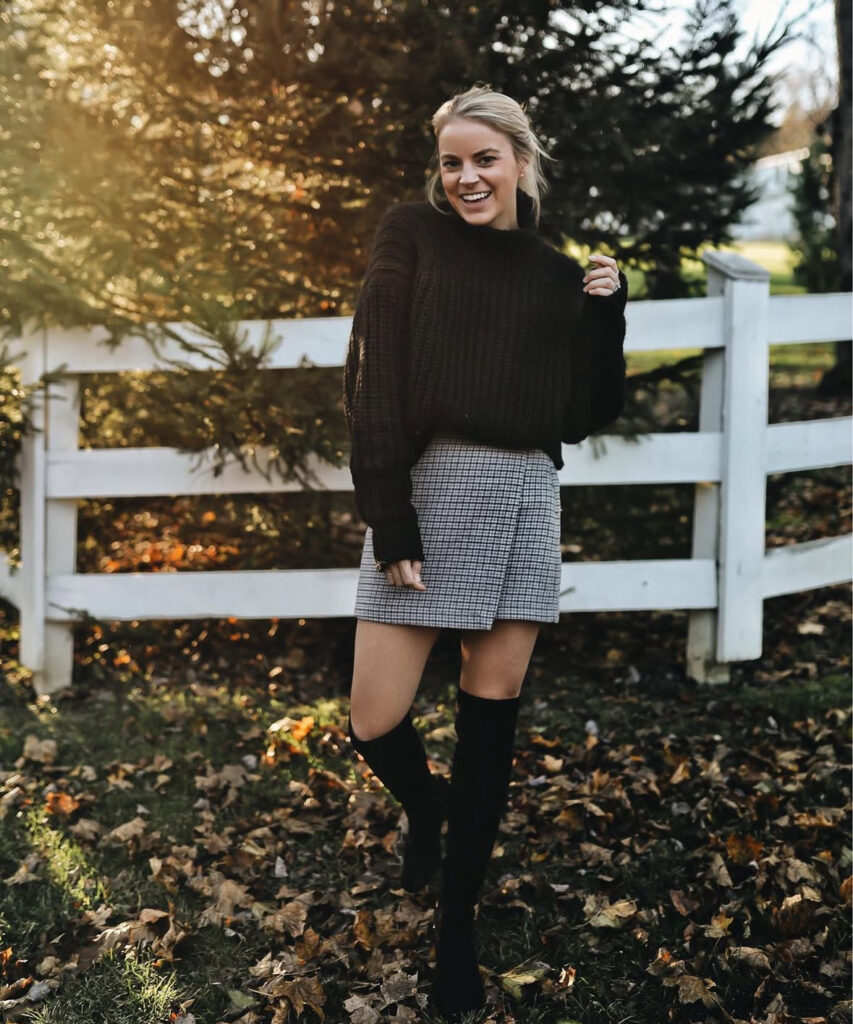 Woman outdoors in a chunky black sweater, gray plaid mini skirt, and black over-the-knee boots near a white fence and fall foliage.