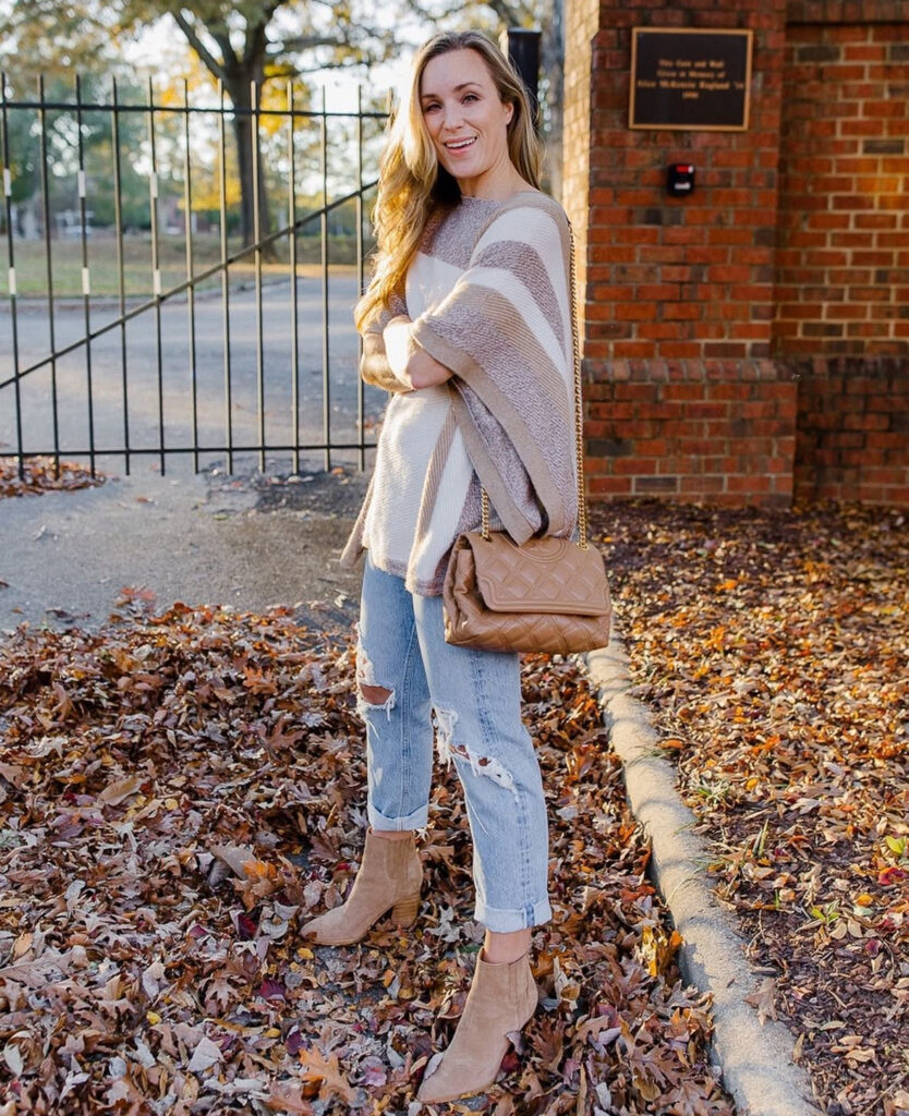 Woman in a striped poncho, distressed cuffed jeans, and tan suede ankle boots with a quilted shoulder bag, standing among autumn leaves.