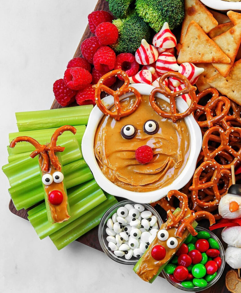 Reindeer-themed snack board with peanut butter dip face, pretzel antlers, candy eyes, raspberry nose, plus celery reindeer sticks and holiday treats.