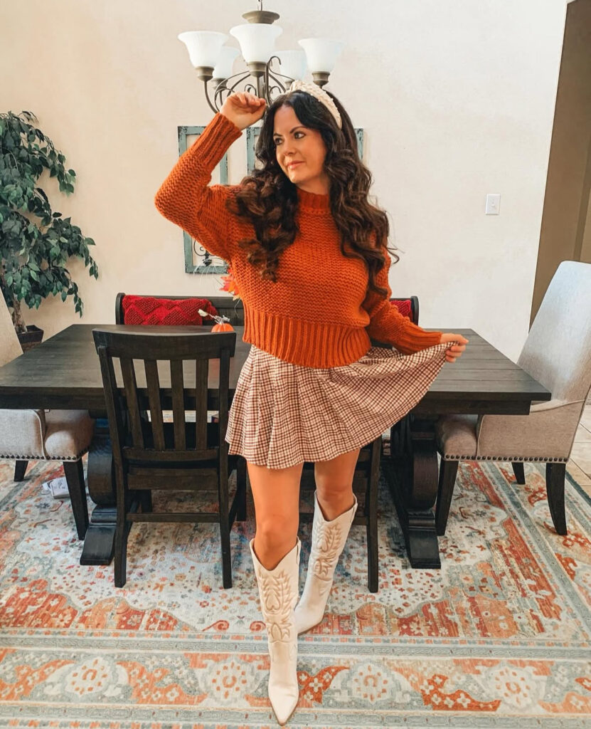 Woman indoors wearing a rust knit sweater, pleated plaid mini skirt, and tall white western boots with a pearl headband.