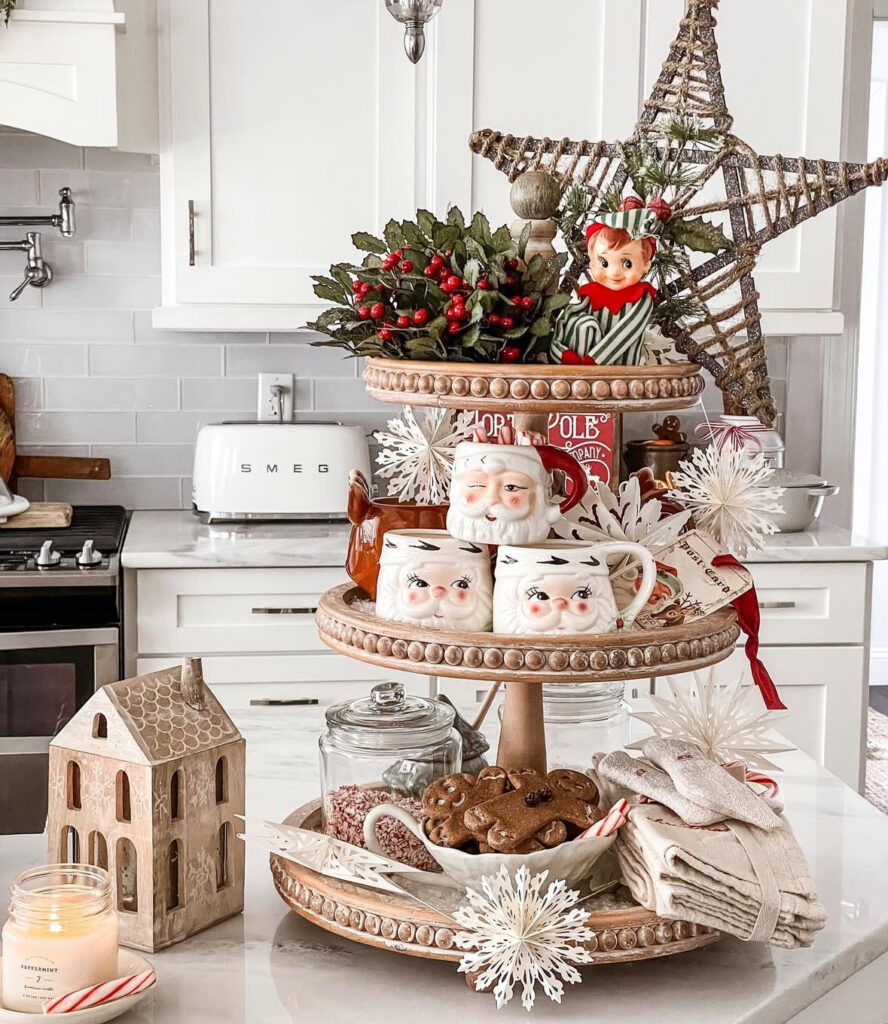 Two-tier tray hot-cocoa station with Santa mugs, elf, cookies, and paper snowflakes on white island.