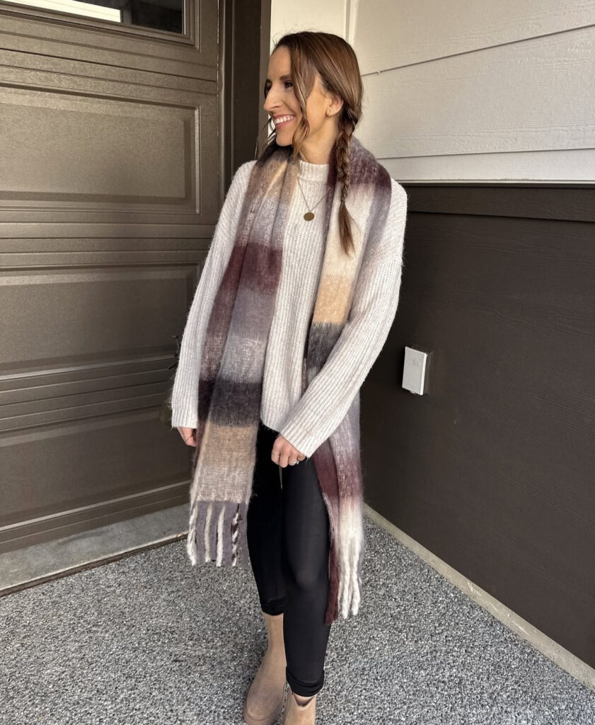 Woman outdoors in a cream ribbed sweater, black leggings, oversized ombré plaid scarf, and tan lug-sole boots, smiling by a garage door.