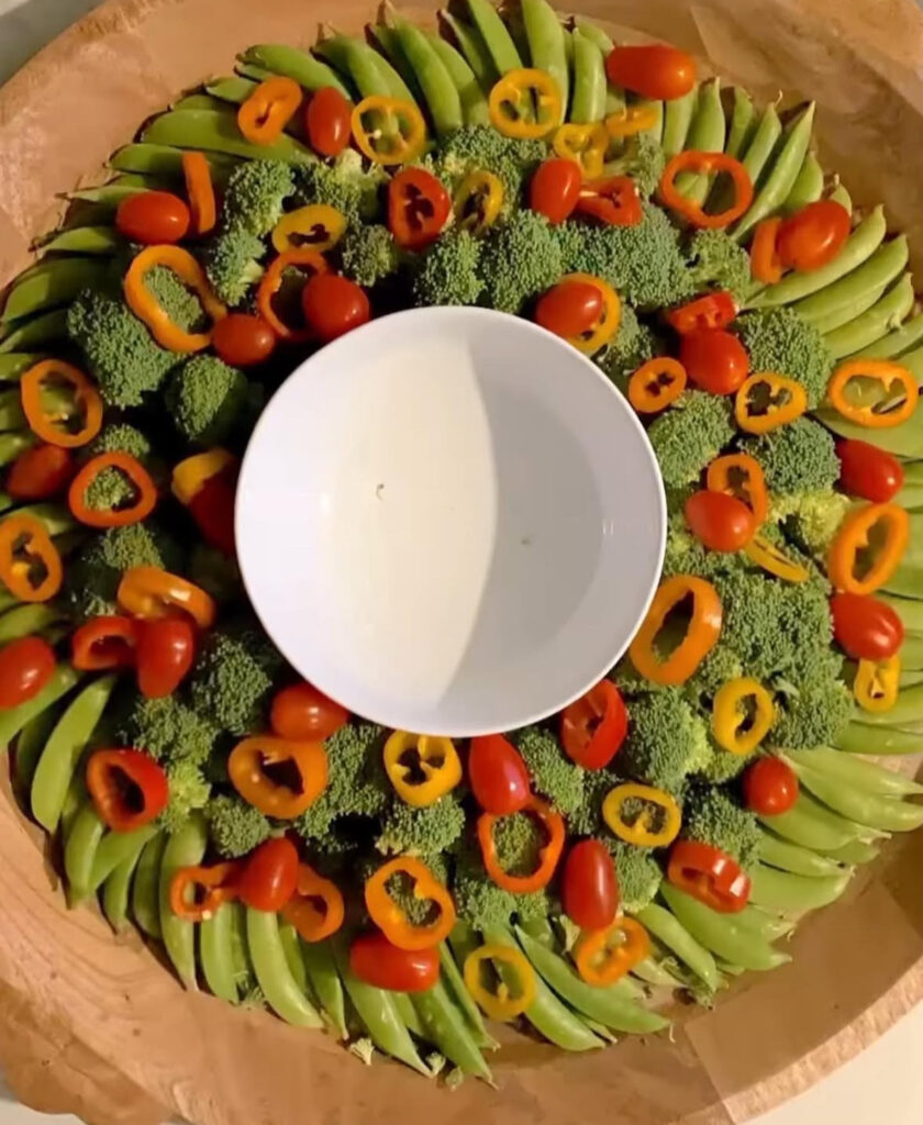 Christmas wreath veggie tray of layered snap peas and broccoli topped with mini pepper rings and grape tomatoes, empty dip bowl in the center.