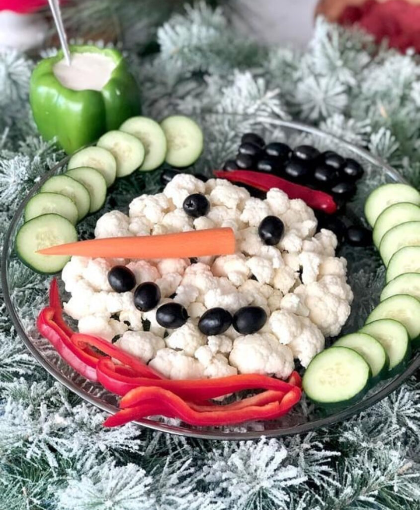 Large snowman platter with three black-lidded bowls as head and body, peppercorn buttons, carrot nose, and a mix of cucumbers, tomatoes, peppers, and broccoli.