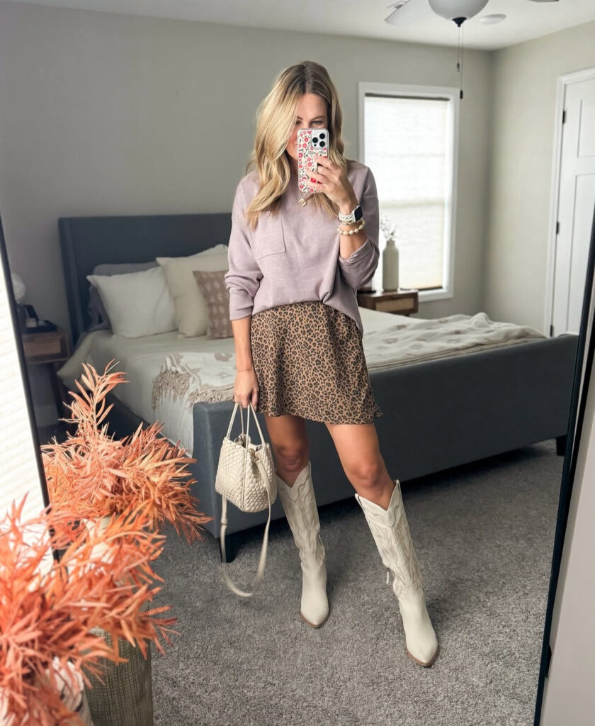 Mirror selfie of lavender pullover with a leopard mini skirt, tall ivory cowboy boots, and a woven bucket bag.