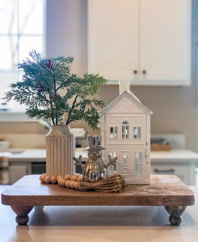 Minimal vignette on wooden riser: ribbed vase of greenery, white house lantern, silver snowman, wood beads.