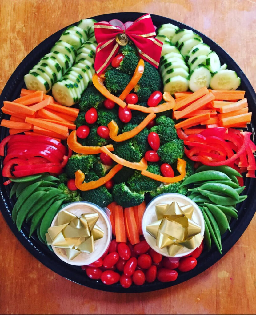 Broccoli Christmas tree platter decorated with tomato “ornaments,” orange pepper “garland,” and a bow on top, flanked by veggies and two dip bowls.